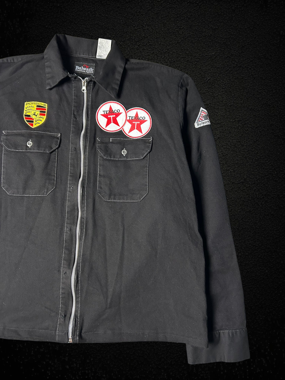 Camino’s Auto Parts reworked black trucker jacket featuring Porsche and Texaco patches with a front zipper closure.